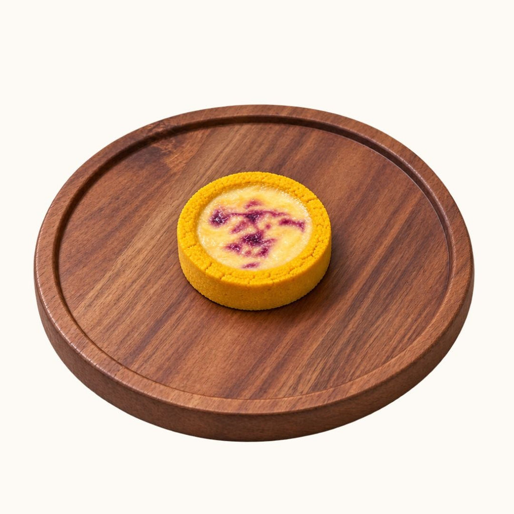 A small round yellow tart or custard pie with a purple berry topping, presented on a round wooden serving board against a plain off-white background.