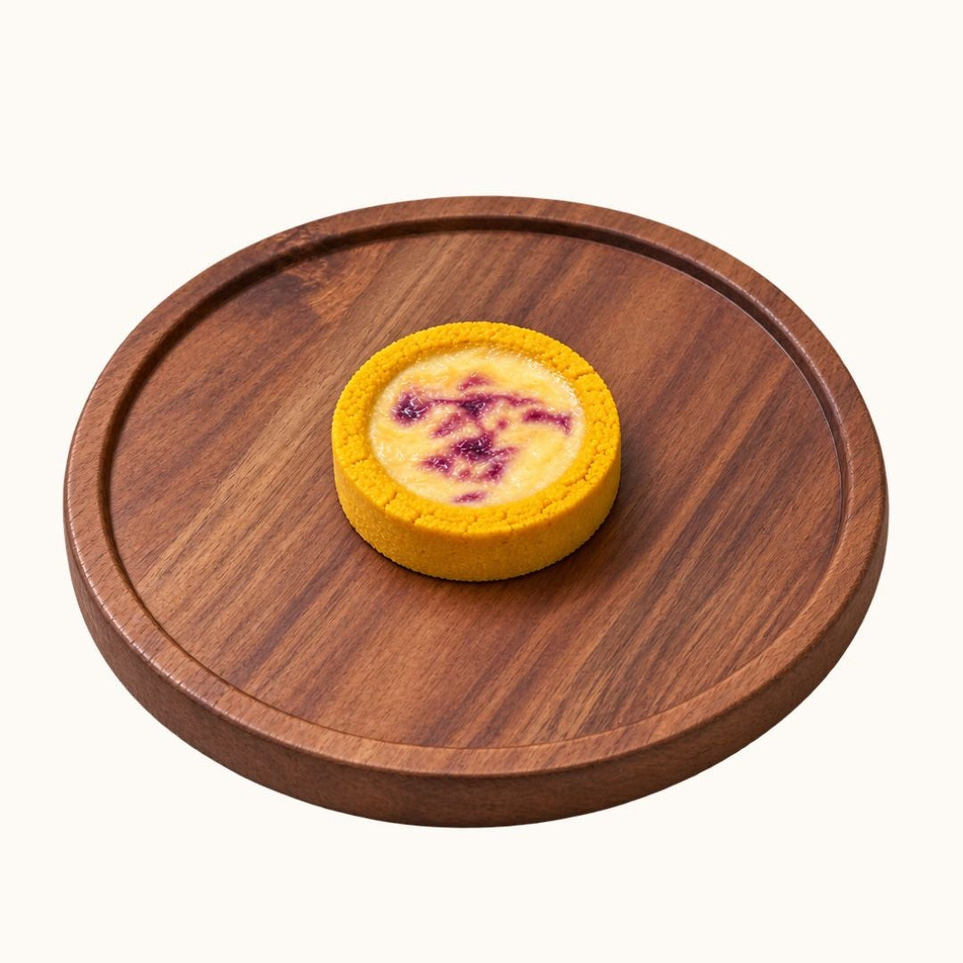 A small round yellow tart or custard pie with a purple berry topping, presented on a round wooden serving board against a plain off-white background.