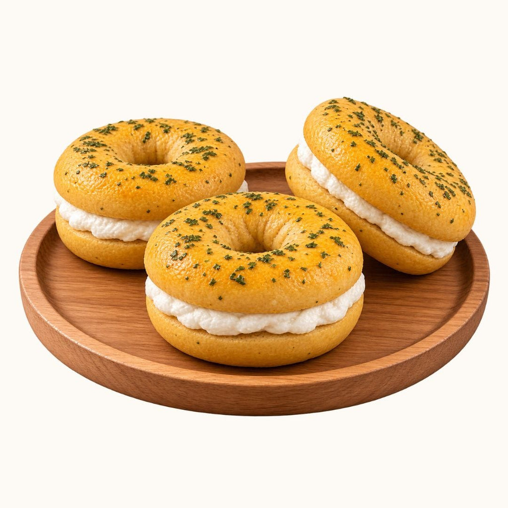 Three bagel halves filled with white cream and topped with green herbs, arranged on a round wooden plate against a plain white background.
