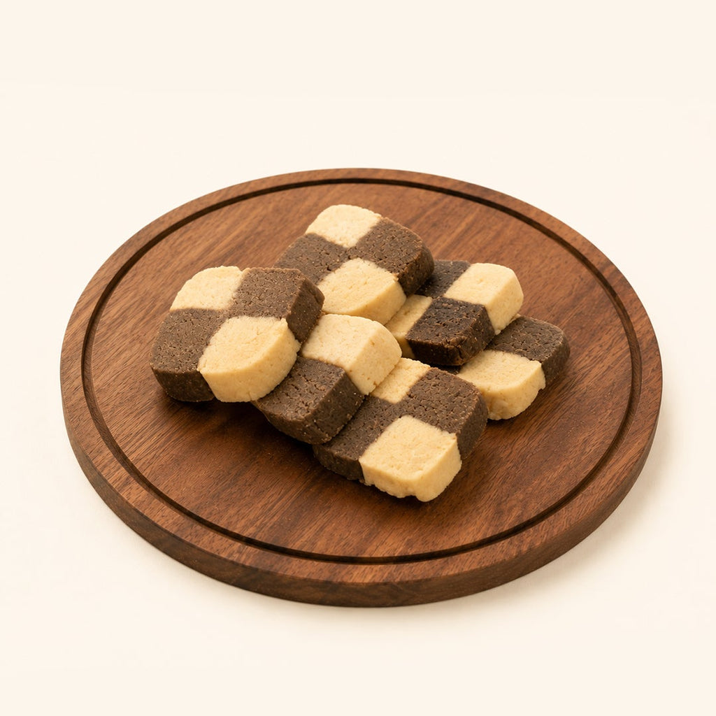 A set of checkered square cookies or candies in dark brown and light beige, arranged on a round wooden serving board against a solid off-white background.