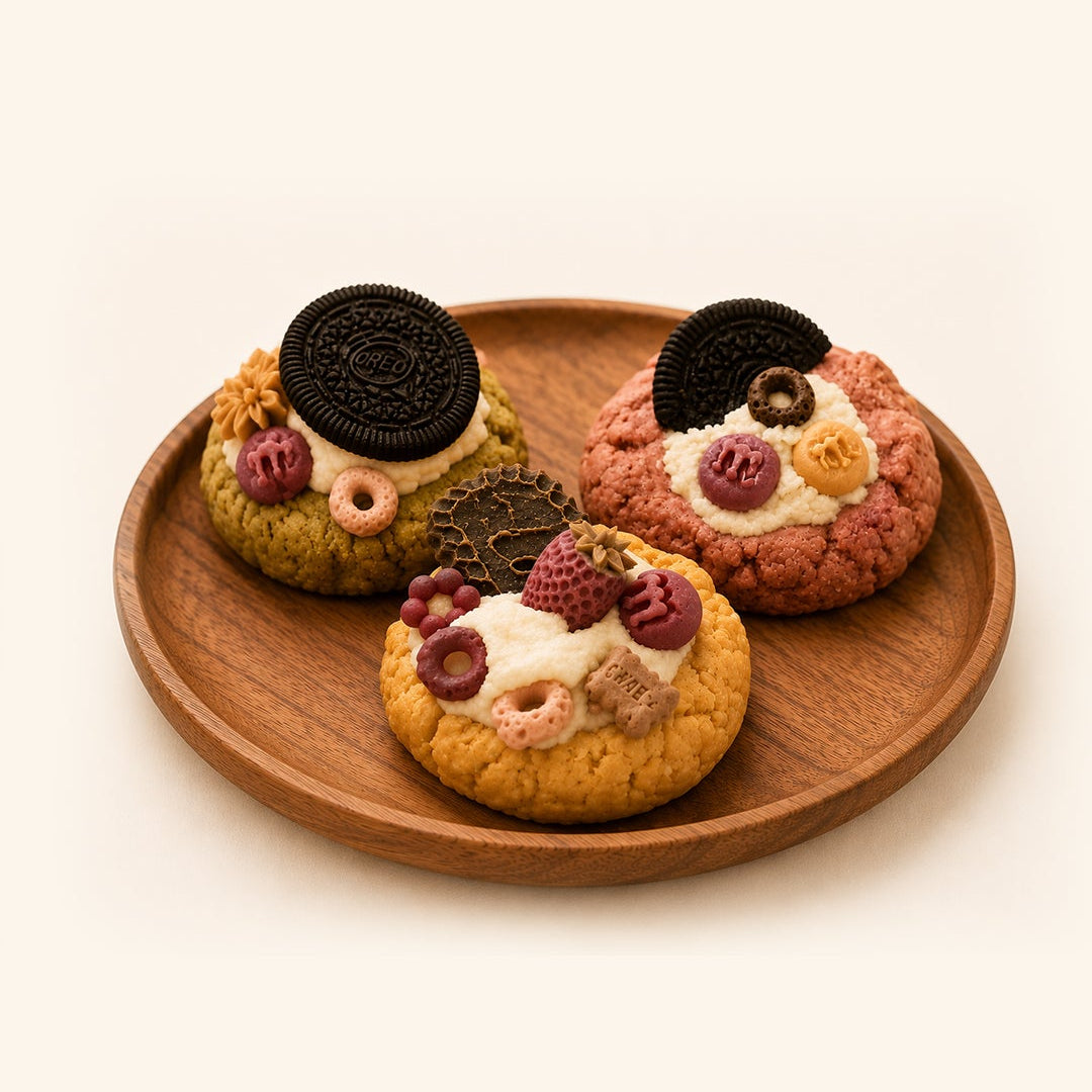 Three decorated cookies arranged on a round wooden plate against a warm off-white background. The cookies have colorful toppings including red berries, pink cream, and dark cookie pieces, with one topped by a whole Oreo cookie.