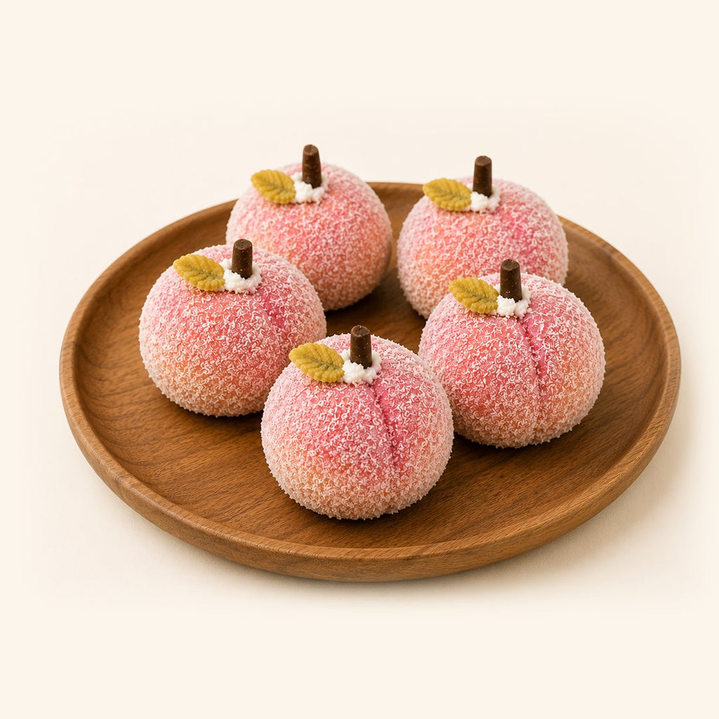 Four peach-shaped sugar-coated candies with small brown stems and yellow-green leaf accents, arranged on a round wooden plate against a warm off-white background.