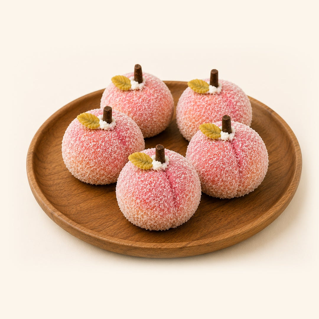 Four peach-shaped sugar-coated candies with small brown stems and yellow-green leaf accents, arranged on a round wooden plate against a warm off-white background.
