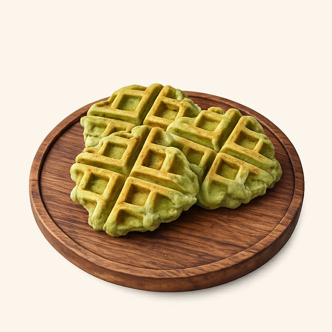 Four green waffles with golden-brown grid lines served on a round wooden serving board against a solid off-white background.